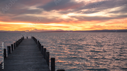 Dusk at Lake Neusiedl with a wooden pier in the left foreground. The turquoise rippled water with an orange reflection streak in the background. The orange red cloudy sky © JürgenBauerPictures