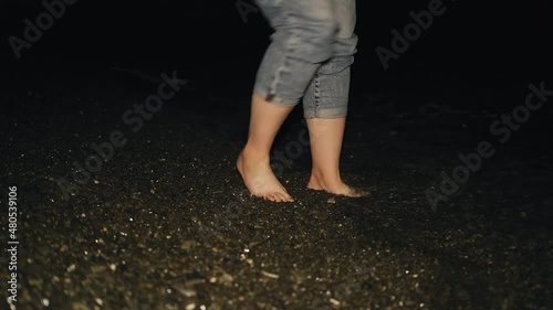 Women's feet at night on a rocky beach. A girl walks by the sea