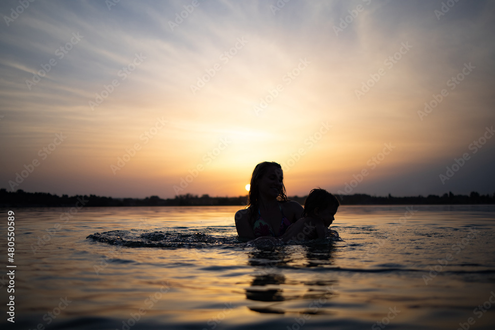 Mom plays with a naked baby in oversleeves in the lake against the background of a summer sunset