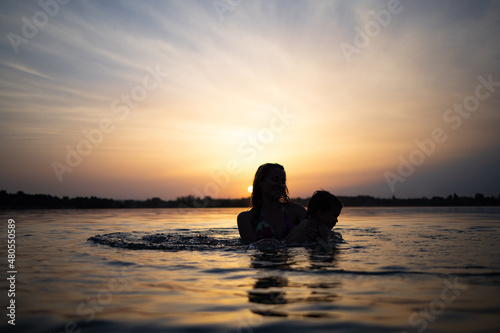 Mom plays with a naked baby in oversleeves in the lake against the background of a summer sunset photo