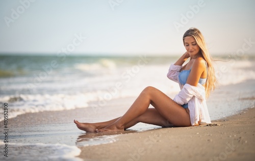 A girl in a bluish swimsuit and shirt sits near the water edge and peers into the horizon © YouraPechkin