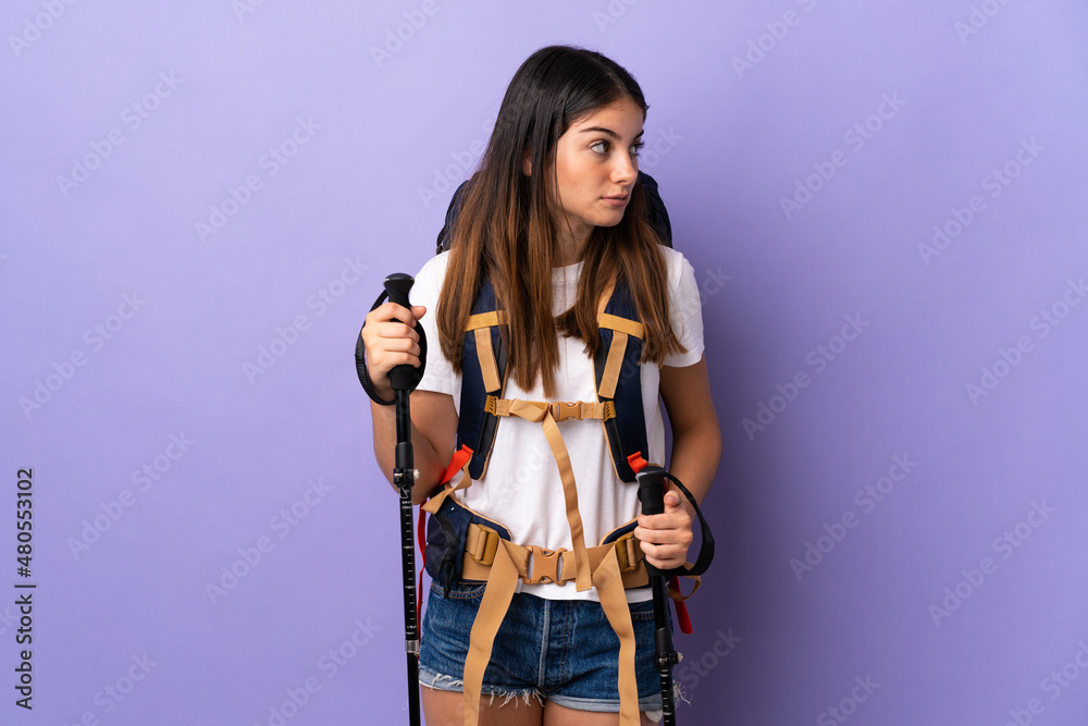Young woman with backpack and trekking poles isolated on purple background looking to the side