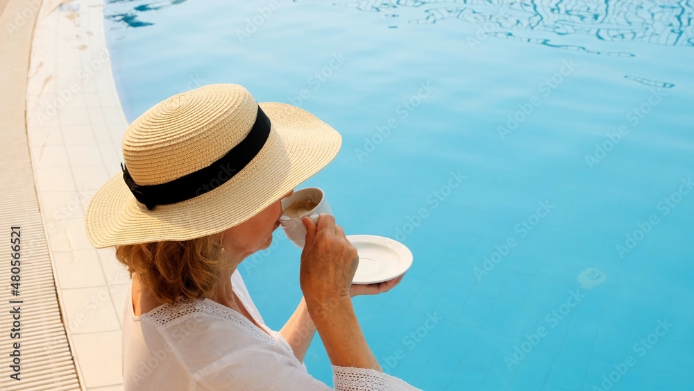Close up of a woman aged 50-55 in a straw hat drinking from a cup of coffee next to a blue swimming pool, luxurious good morning, start of the day