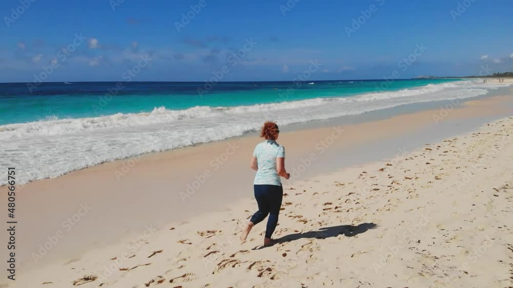 SLOW MOTION: Back view woman running at seashore with azure water at sunny day and bright sunshine slow motion. Adult plump woman runs barefoot along coast of sea on sand. Healthy lifestyle by the sea SLOW MOTION: Back view woman running at seashore with azure water at sunny day and bright sunshine slow motion. Adult plump woman runs barefoot along coast of sea on sand. Healthy lifestyle by the sea