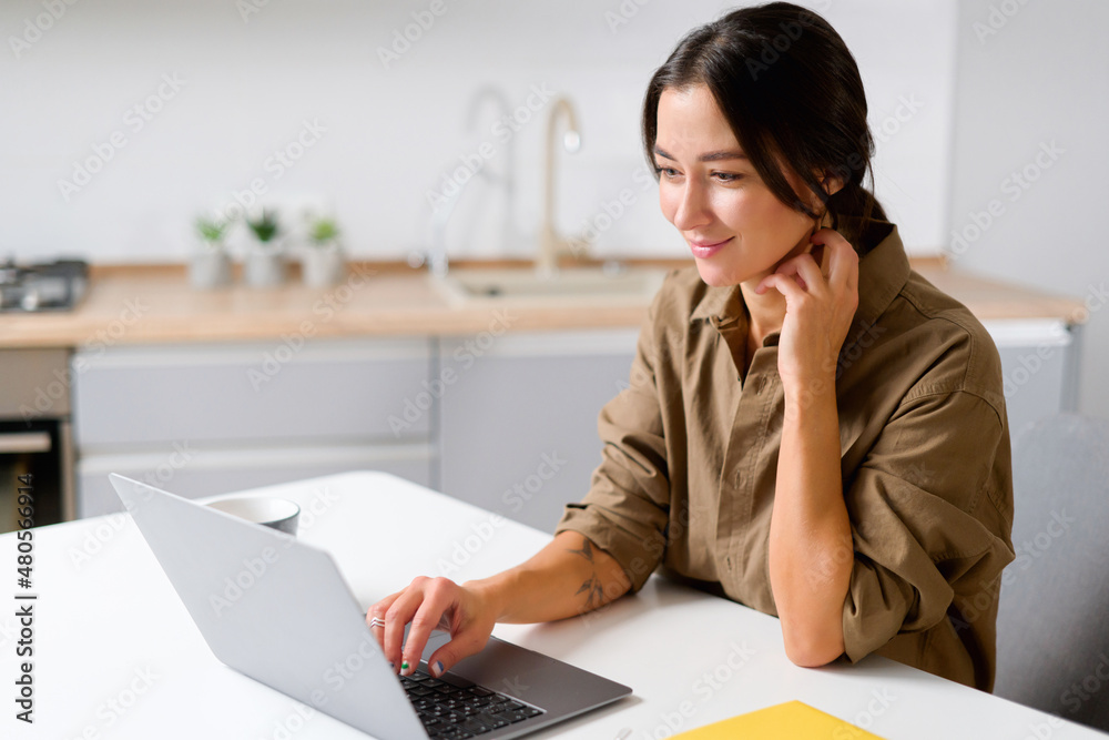 Cheerful asian girl using laptop for remote work or home leisure while sitting in the table at home. Side view of the nice girl looking at the screen with smile Cheerful asian girl using laptop for remote work or home leisure while sitting in the table at home. Side view of the nice girl looking at the screen with smile