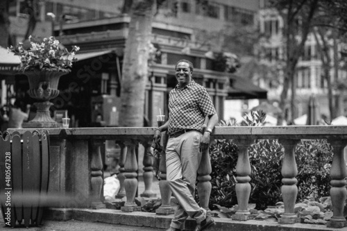 Fashionable portrait of middle aged elegant afro American man wearing brown suit walking in park near to fence in New York. Smiling and positive. Black and white photo