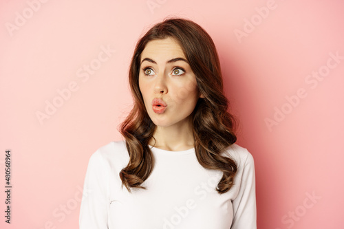 Image of stylish glamour woman looking surprised and amazed, saying wow with impressed reaction, standing over pink background