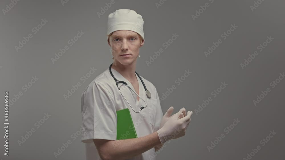 Young Doctor In Medical Gown And Gloves. He Holds Notebook Under His Arm. Young Doctor In Medical Gown And Gloves. He Holds Notebook Under His Arm.