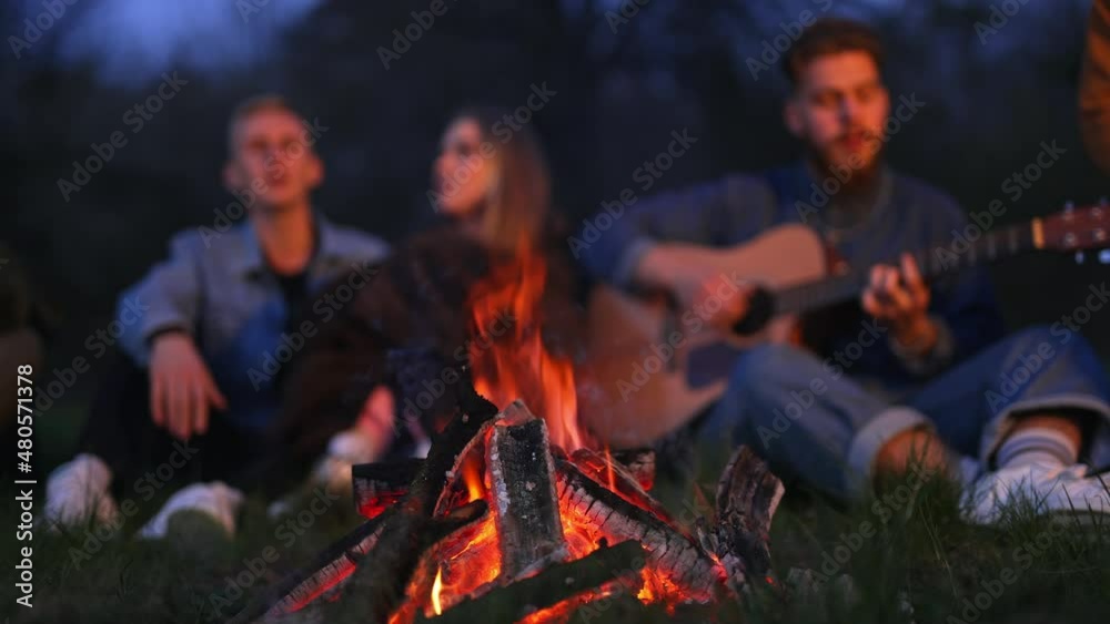 Cozy bonfire burning at nighttime. Friendly campers singing song along with the guitar at the backdrop in blur. Cozy bonfire burning at nighttime. Friendly campers singing song along with the guitar at the backdrop in blur.