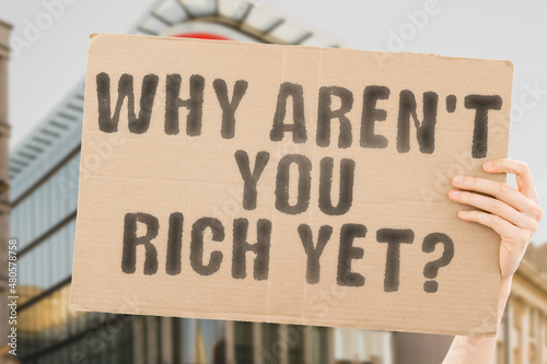 The question " Why aren't you rich yet? " on a banner in men's hand with blurred background. Planning. Payment. Save. Win. Entrepreneurship. Work. Persistence. Million. Accumulate