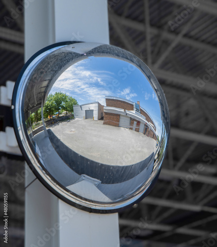 Large convex safety and security mirror mounted on a white steel beam, reflecting a roadway and garage buildings, daytime, nobody.