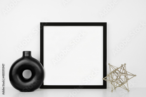 Mock up black square frame with modern home decor. White shelf against a white wall. Circle vase and geometric object. Copy space.