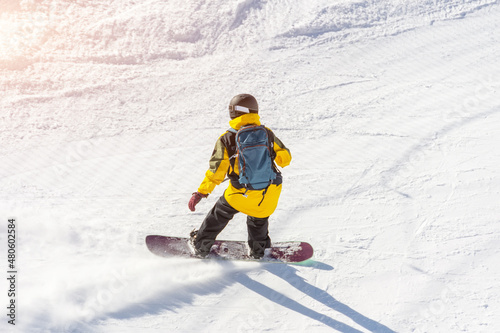 Snowboarder in a helmet and with a backpack on his back rides along a mountain track on a sunny day.