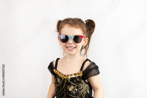 A little playful girl with two ponytails and a black and gold dress. Studio photo on a white background of a child in sunglasses © PeterPike
