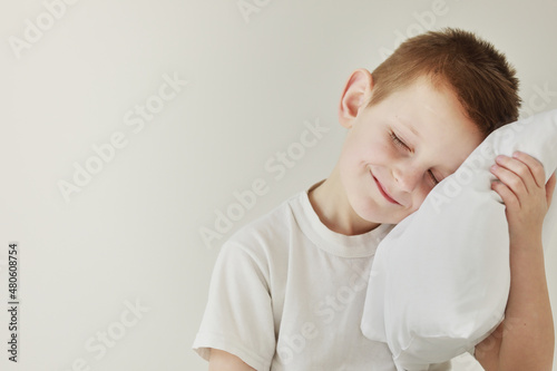 Portrait of casually dressed little boy leaning on white pillow, while hugging it, feeling tired after long full day, with cheerful smile, having rest
