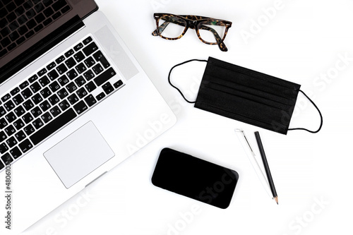 Laptop keyboard, medical face mask, reading glasses, notebook, pen and mobile phone on white office desk. New normal at work