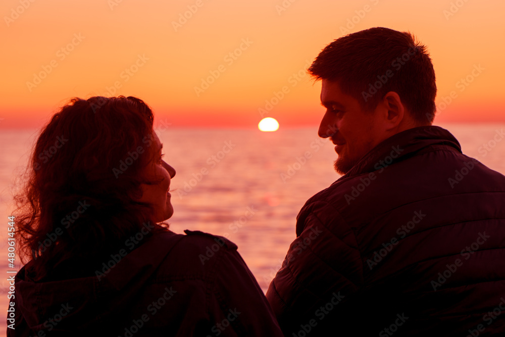 Travelers. Husband and wife. Date. Romance. A day off in nature. A man and a woman sit on the stone beach and enjoy the winter sea and the beautiful orange bright sunset
