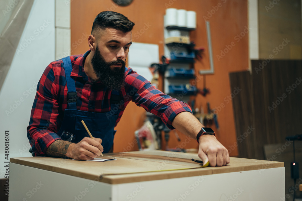 Young male carpenter working in workshop and measuring with meter Young male carpenter working in workshop and measuring with meter