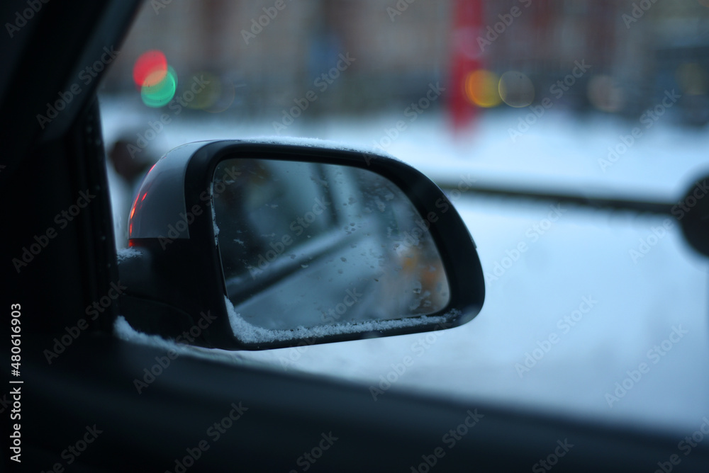 Snow covered car rear view mirror on bokeh winter cityscape background.