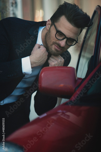 Handsome gentleman in suit and with bow tie,shirt, with glasses, looking at car mirror