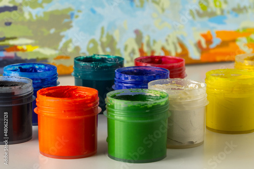 Open jars of multi-colored gouache on a blurred watercolor background. The concept of learning to draw. Materials for creativity.