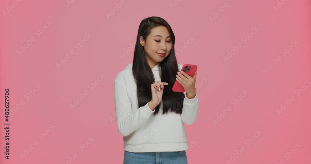 Funny pretty young asian girl playing online game or chatting using modern mobile phone application on pink background Funny pretty young asian girl playing online game or chatting using modern mobile phone application on pink background