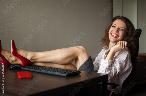 Portriat of brunette with legs on office table with laptop, hot woman in red shoes on high heels sitting at the desk. Concept of seductive secretary, female fashion, relax at work