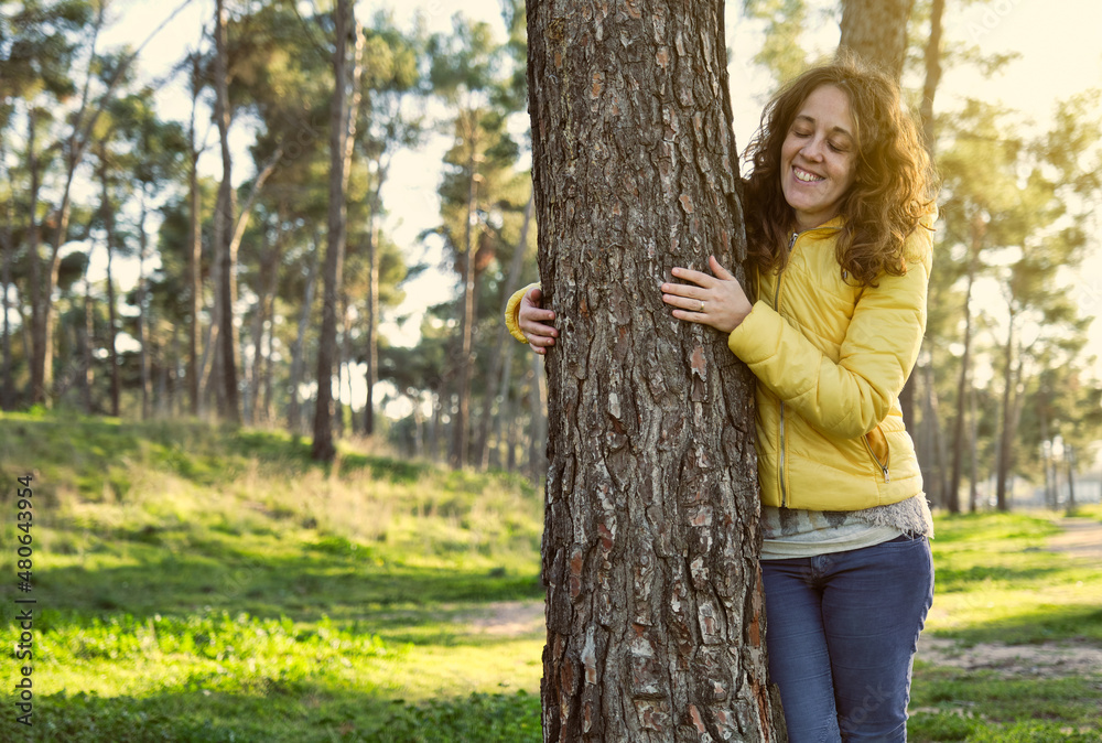 young girl smiling with closed eyes wearing yellow jacket and blue pants from the front hugging a tree in a pine forest at sunset young girl smiling with closed eyes wearing yellow jacket and blue pants from the front hugging a tree in a pine forest at sunset