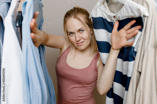  Happy young white woman looks out from behind clothes hanging on a hanger, smiles at the camera, is satisfied with the choice of purchases, buys a summer wardrobe in bed color