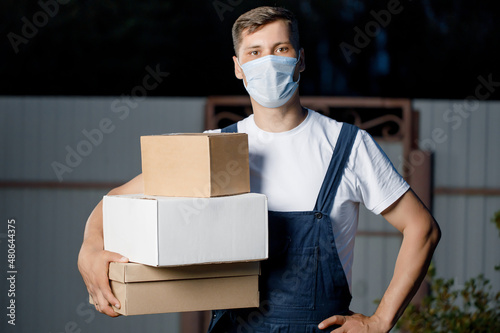 Courier man in uniform and mask holds a stack of boxes in his hands. Courier delivery service with parcels in hand in the open air