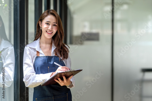 Beautiful young Asian woman entrepreneur wear an apron standing taking note looking at camera beside a large mirror.