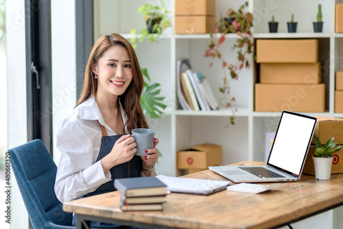 Asian business owner selling products online sitting holding a cup of coffee with a laptop computer smiling happily at desk office.