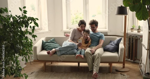 Couple and 5s daughter spend time together sit on sofa with cellphone use new mobile app, having fun enjoy internet amusing content enjoy carefree weekend. Family leisure at home, modern tech concept