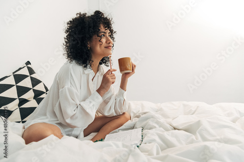 Valokuva Young mixed race woman sitting on bed having coffee and writing on her journal