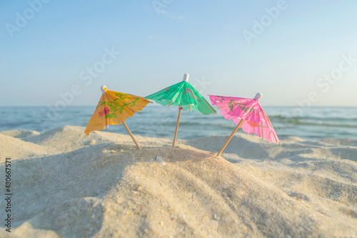 Three small paper cocktail umbrellas stand in sand on sandy beach close-up.