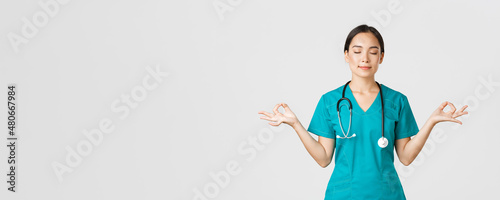 Covid-19, healthcare workers and preventing virus concept. Calm and patient asian female doctor, nurse in scrubs staying relaxed, meditating with eyes closed and happy smile, white background