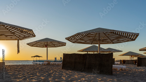 There is no one on the Red Sea beach in the early morning. The sun is shining above the horizon. Lattice sun umbrellas against the blue sky. Light and shadows on the undulating sand. Egypt