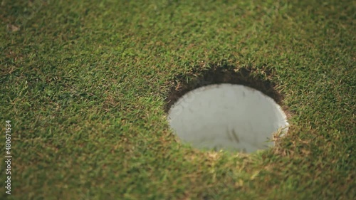 A white golf ball rolls right into hole on a golf course with green grass, close-up. Moment when the white golf ball hits the hole. The moment of playing golf. The exact hit of the ball on target.