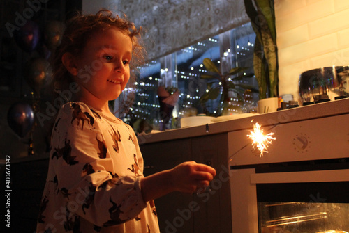 A little girl with curly hair holds a sparkler in her hand. The girl is happy, she has a holiday in a homely cozy atmosphere, she looks with admiration at the sparkler in her hand.