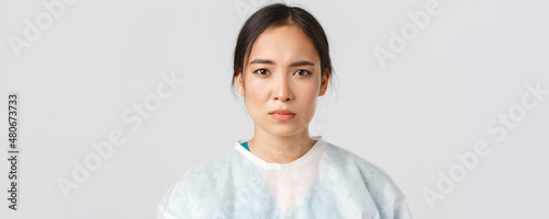 Covid-19, coronavirus disease, healthcare workers concept. Serious exhausted asian female doctor looking tired after shift at hospital, take-off personal protective equipment, have skin marks photo