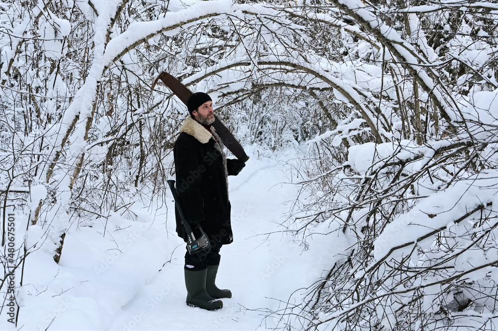 brutal man with beard and warm clothes is standing in middle of snowy forest. Logger holds an axe and hand saw. Road and trees are covered with snow. Far from civilization. Harvesting of firewood