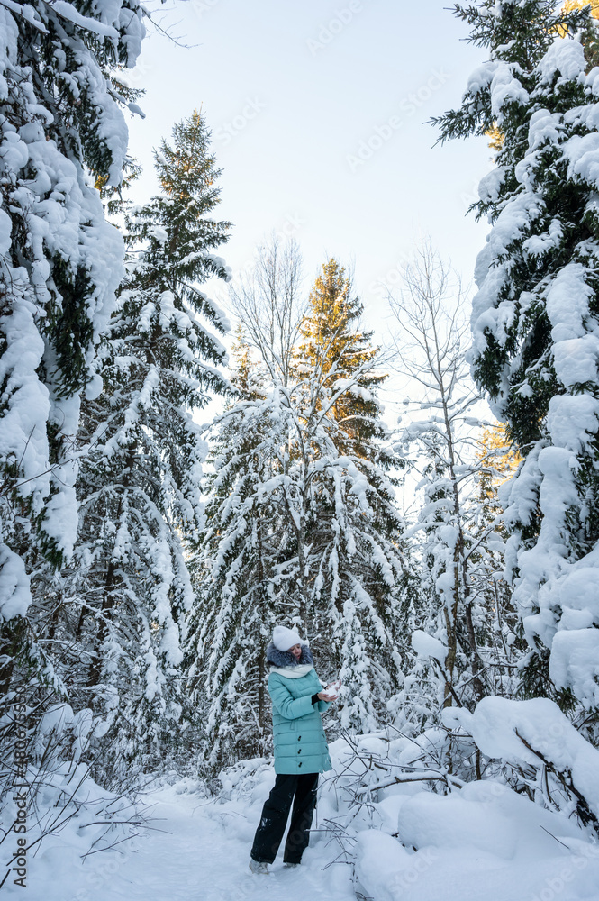 young girl walks in winter forest among tall fir trees covered with snow. Girl is wearing warm jacket, white hat and scarf. Clothes for walking in winte