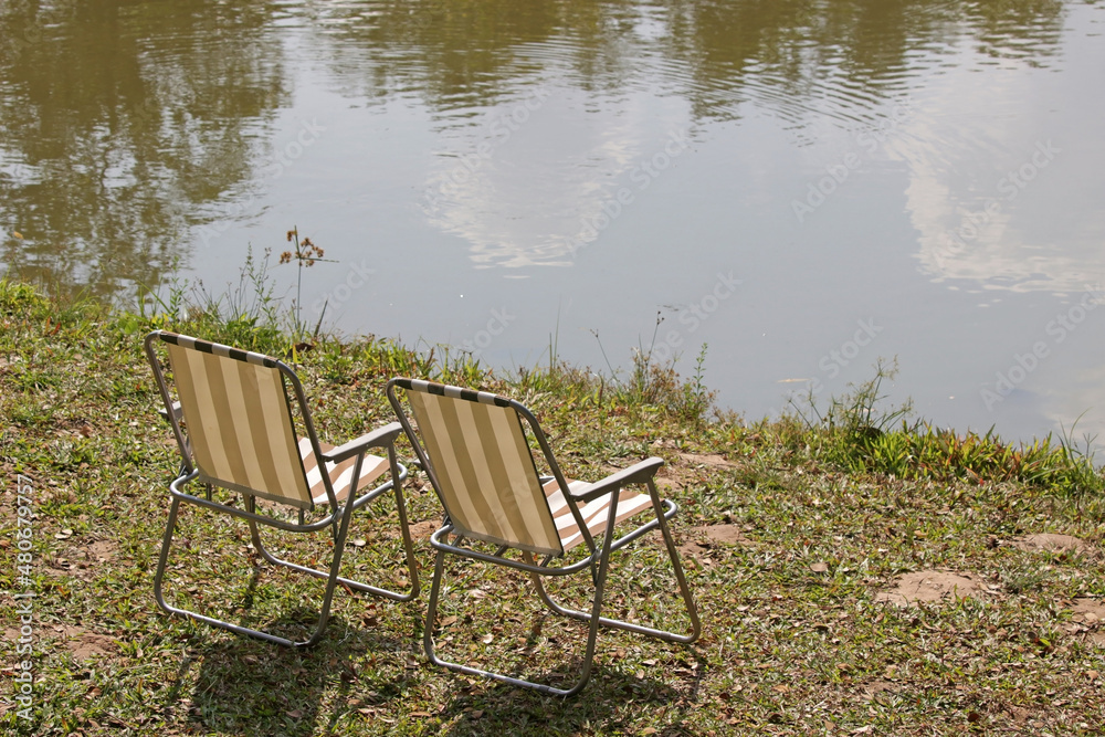 Fototapeta premium Pair of empty folding garden chairs on grass next to the edge of a river or a lake on a sunny summer day. No people.