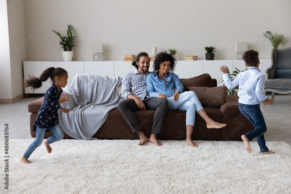 Happy positive Black couple of parents watching two energetic hyperactive little kids running around in living room, chasing, playing active games, having fun. Parenthood, upbringing, family concept Happy positive Black couple of parents watching two energetic hyperactive little kids running around in living room, chasing, playing active games, having fun. Parenthood, upbringing, family concept