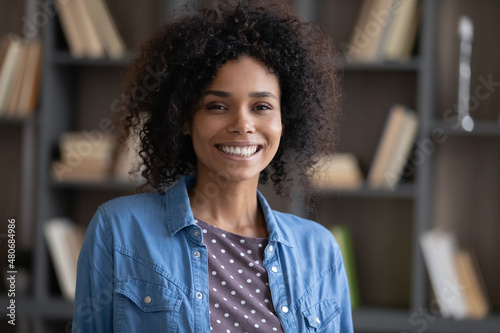 Happy beautiful curly haired millennial Afro American woman in casual home head shot portrait. African girl posing in apartment interior with toothy smile, laughing, looking at camera