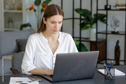 Young focused caucasian businesswoman sitting at desk working on a laptop computer in contemporary office