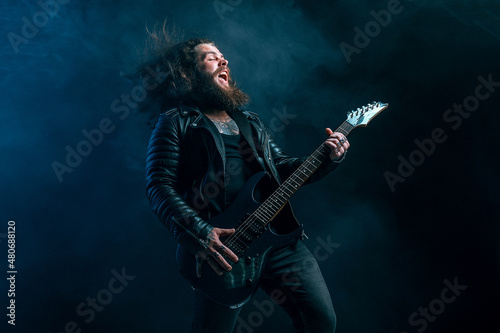 Expressive rock male guitarist with long hair and beard plays on the smoke background. Studio shot