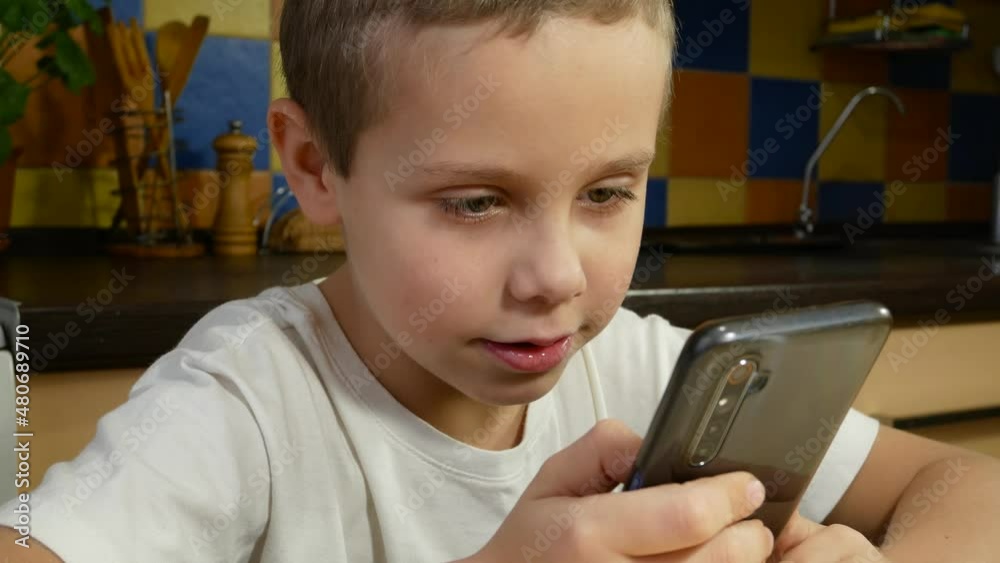 A cute boy of 6-7 years old smiles and talks, looking at the screen of a smartphone. Scroll the screen with your finger. Communication and learning using a smartphone A cute boy of 6-7 years old smiles and talks, looking at the screen of a smartphone. Scroll the screen with your finger. Communication and learning using a smartphone