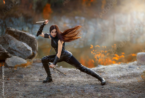 Fantasy fighting woman assassin actions in motion battle, hold daggers in hand. Red-haired girl warrior in black leather costume. ninja soldier with knives. Red long hair fluttering fly in wind