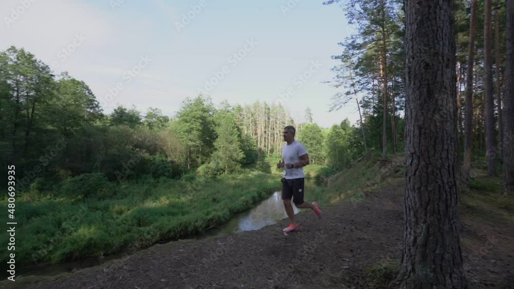 adult man in sportswear runs along edge of cliff near river in forest area on sunny day